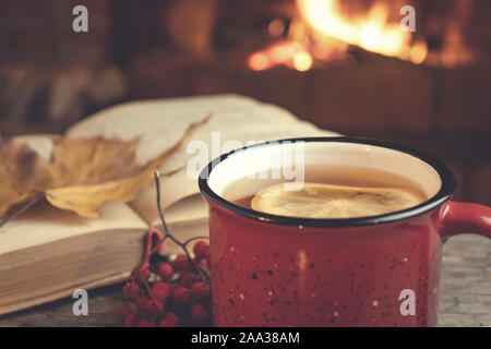 Mug rouge avec du thé chaud et un livre ouvert en face d'une cheminée à bois, confort, détente et chaleur de l'âtre concept. Banque D'Images