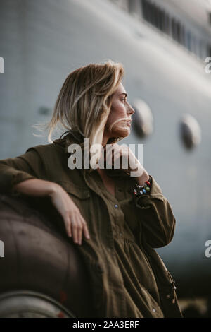 Belle femme blonde en manteau vert, debout près de l'ancien avion volant. Banque D'Images