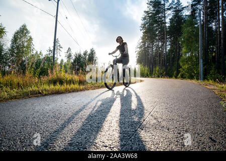 Happy woman rides un vélo sur une piste cyclable à proximité d'une forêt de pins. Elle sourit et profitant de la pluie d'été chaude et le soleil. Banque D'Images