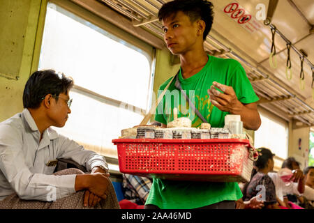 Birman jeune vendeur vend ses marchandises vers le bas-côté d'un train à partir d'un panier en plastique tout en montant le train qui encercle le centre de Yangon, Myanmar (Birmanie) Banque D'Images