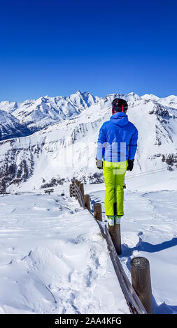 L'homme dans une tenue de ski debout sur un pilier en bois et profiter de la vue du Alp. Homme porte veste bleue et pantalon vert fluo. Le plein de bons powd Banque D'Images