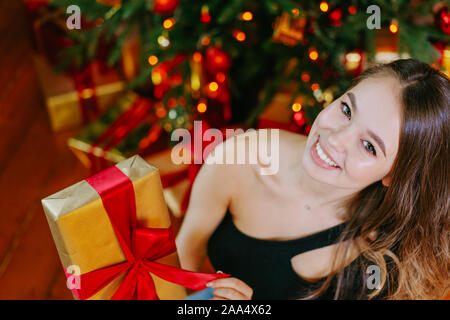 Belle jeune fille tenir don et sourire, assis près de l'arbre de Noël à la maison. Don de l'or avec red bow Banque D'Images