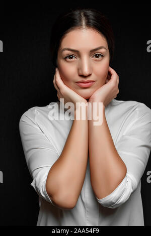 Portrait d'une femme brunette asiatique dans une chemise blanche couvrant son visage et les oreilles avec sa main sur un fond noir Banque D'Images