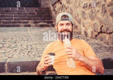 Ce qu'un casse-croûte. Homme barbu malsain de manger sandwich hot dog snack. Le repos et le Hipster ayant hotdog snack sur escalier extérieur. Guy caucasienne bénéficiant d'un casse-croûte sur rue. Banque D'Images