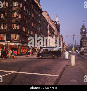 Années 1960, historique, policier dans un manteau blanc qui se tenait sur le chemin de diriger un van noir de l'époque dans la rue Princes Street, Edinburgh, Ecosse. Au coin de South St David Street, on peut voir le magasin de détail de Dunn & Co et de l'hôtel Old Waverley, ainsi qu'un autobus routemaster rouge dans la distance. Banque D'Images