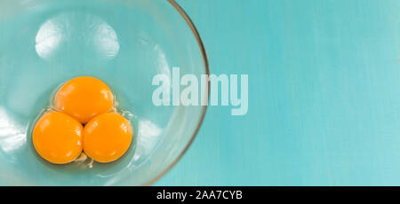 Trois matières premières fraîches d'œufs dans un bol en verre préparé pour fouetter sur fond bleu. Banque D'Images