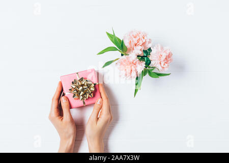 Woman's hands holding boîte cadeau rose à côté de petit bouquet de fleurs sur la table en bois blanc, vue du dessus. Banque D'Images