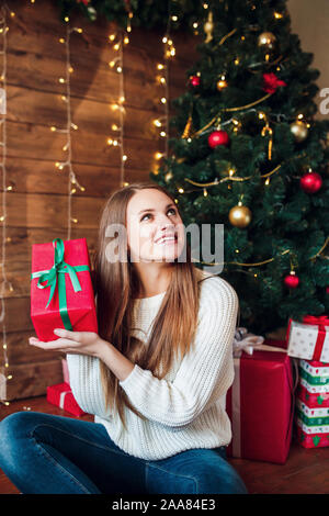 Beautiful Girl holding a Christmas present . Happy woman in Santa hat assis près de Nouvel An des arbres Banque D'Images