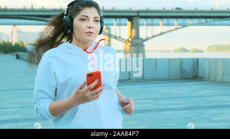 Belle fille dans un chandail blanc et un casque sur un matin dans le jogging. Elle court le long de la promenade et écoute de la musique Banque D'Images