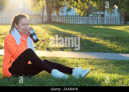 Belle jeune femme dans un chandail sport rouge est assis sur l'herbe après un jogging. Elle boit de l'eau d'une bouteille en métal Banque D'Images