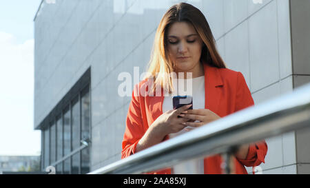 Business Woman in a Red Jacket lit un message dans un smartphone, se trouve à proximité d'un centre d'affaires Banque D'Images