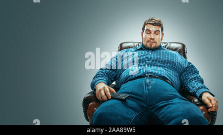 Portrait de fat man wearing jeanse et whirt assis dans un fauteuil brown gradient isolé sur fond gris. Regarder la télévision et émotionnel d'un changement de chaîne, laughting. Surpoids, sans soucis. Banque D'Images