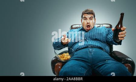 Portrait de fat man wearing jeanse et whirt assis dans un fauteuil brown gradient isolé sur fond gris. Regardez la télévision boit de la bière, mange des frites et des canaux en évolution. Surpoids, sans soucis. Banque D'Images