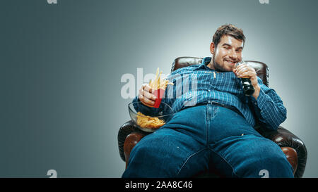 Portrait de fat man wearing jeanse et whirt assis dans un fauteuil marron dégradé sur fond gris. En regardant la télé, mange des boissons cola, chips de pommes de terre frites, laughting. Surpoids, sans soucis. Banque D'Images