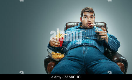 Portrait de fat man wearing jeanse et whirt assis dans un fauteuil marron dégradé sur fond gris. En regardant la télé, mange des boissons cola, chips de pommes de terre frites, laughting. Surpoids, sans soucis. Banque D'Images