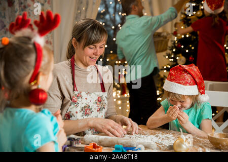 Mère et enfants en bonnets rouges la cuisson gingerbread cookies et de décoration de la glaçure. Père de l'enfant avec la décoration de sapin de Noël. Belle salle de séjour Banque D'Images