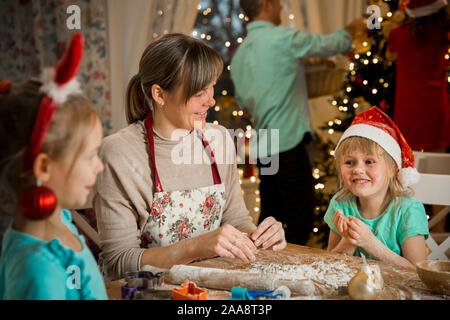 Mère et enfants en bonnets rouges la cuisson gingerbread cookies et de décoration de la glaçure. Père de l'enfant avec la décoration de sapin de Noël. Belle salle de séjour Banque D'Images