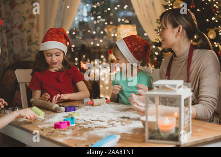 La mère et les petits enfants en bonnets rouges la cuisson gingerbread cookies et de jouer. Beau salon avec des lumières et l'arbre de Noël, une table avec la lanterne. Banque D'Images