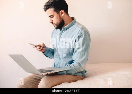 Man holding laptop et smart phone while sitting on bed at home Banque D'Images