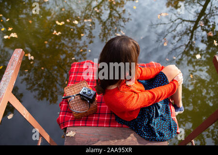 Une jeune femme assise avec son dos sur un pont près d'un lac sur une journée ensoleillée. Plaid, appareil photo, suitcase Banque D'Images