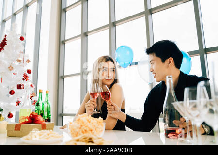 Young adult Asian couple tenant un verre de vin célèbre la fête de Noël au condo de luxe avec des collations, des cadeaux, arbre de Noël sur la table. Banque D'Images