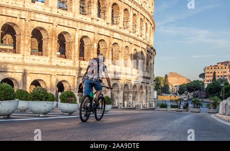 Heureux jeune homme avec le vélo touristique portant chemise et hat au Colisée à Rome, Italie au lever du soleil. Banque D'Images