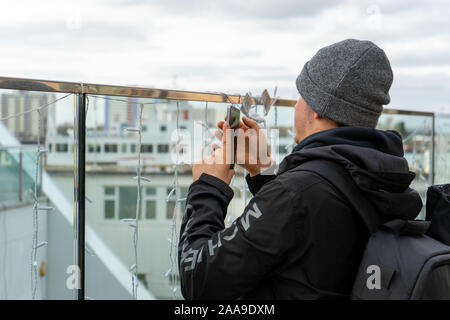 Un homme dans la vingtaine, portant un chapeau de laine à l'aide d'un téléphone mobile ou téléphone cellulaire pour prendre une photo d'une attraction touristique Banque D'Images