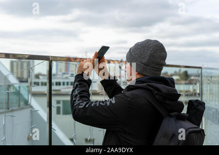 Un homme dans la vingtaine, portant un chapeau de laine à l'aide d'un téléphone mobile ou téléphone cellulaire pour prendre une photo d'une attraction touristique Banque D'Images