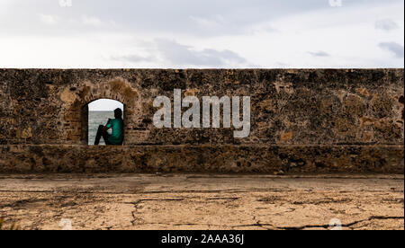 Avec l'homme en chemise verte assis sur le mur de Carthagène, en regardant son téléphone cellulaire avec la mer en arrière-plan et le plancher à l'avant-plan Banque D'Images