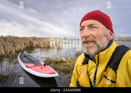 Portrait de l'environnement concernés et un concentré de pagayeur senior gilet avec son paddleboard et le lac en arrière-plan, un paysage de printemps précoce wit Banque D'Images