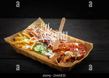 Boîte en carton rempli de nourriture barbecue : kebab, légumes, frites et sauce mayonnaise sur fond noir. Banque D'Images