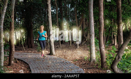 Senior woman running jogging exerçant le long d'un chemin à la tombée de la nuit avec la lumière à travers les arbres Banque D'Images