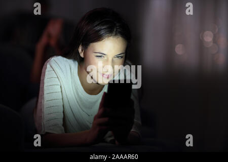 Jeune fille sérieuse à l'aide de smart phone dans la nuit avec le visage allumé allongé sur un canapé dans la nuit à la maison Banque D'Images