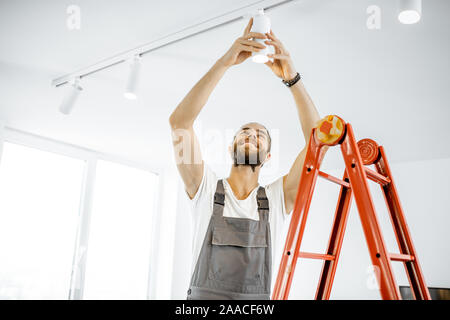 Réparateur ou électricien en vêtements de l'installation de taches claires, debout sur l'échelle dans le salon blanc Banque D'Images