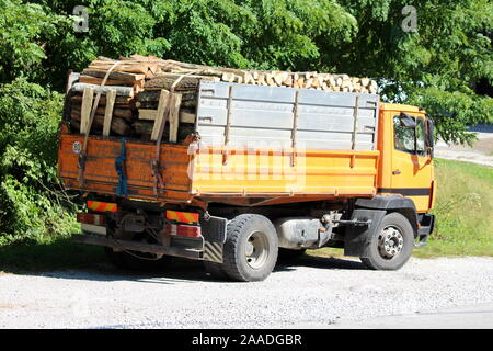 Vue arrière de l'ancien camion rempli de bois jaune garée sur parking de gravier à côté de route pavée entourée d'arbres denses sur l'été chaud et ensoleillé Banque D'Images