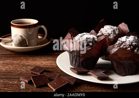 Nature morte avec une tasse de thé ou café et muffins au chocolat trois saupoudré de sucre. Plaque blanche sur tableau noir haut. L'espace pour votre texte, noir backg Banque D'Images