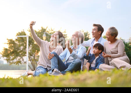 Trois génération happy asian family sitting on grass en tenant un téléphone mobile utilisant selfies en plein air park Banque D'Images