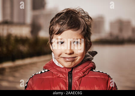 Un garçon de sept ans, dans des vêtements chauds, se dresse sur une rue de la ville dans le froid et nuageux avec un sourire sur son visage. Portrait d'un enfant gai dans Banque D'Images