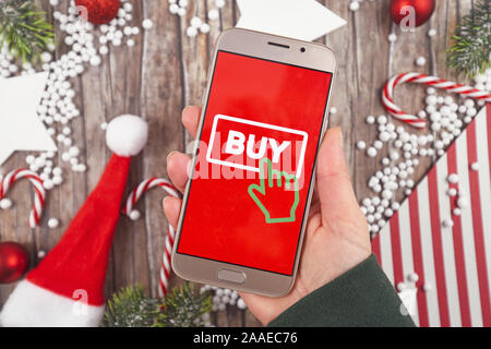 Concept pour la saison de Noël le magasinage en ligne et des ventes avec hand holding cell phone avec le bouton 'Acheter' signe en arrière-plan rouge en face de bureau en bois Banque D'Images