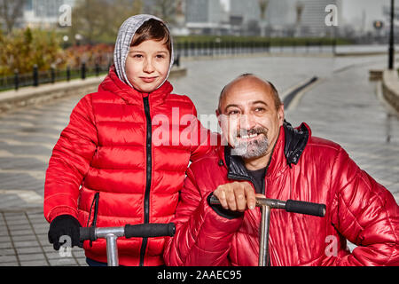 Père et fils ride scooters par temps froid, un garçon de 7 ans et l'homme d'âge moyen vêtus de vêtements chauds à pied le long d'une rue de la ville sous la pluie. Une fam Banque D'Images