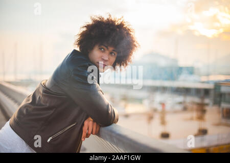 Jeune homme de race mixte avec afro hairstyle debout à une main courante avec beau coucher de soleil avec l'arrière-plan urbain Banque D'Images