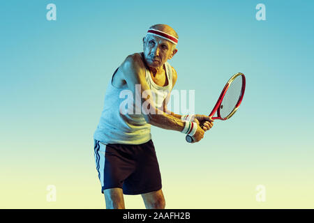 Senior man wearing sportwear jouer au tennis sur fond dégradé, neon light. Modèle homme de race blanche dans la grande forme reste actif, sportif. Concept de sport, activité, mouvement, bien-être, la confiance. Banque D'Images