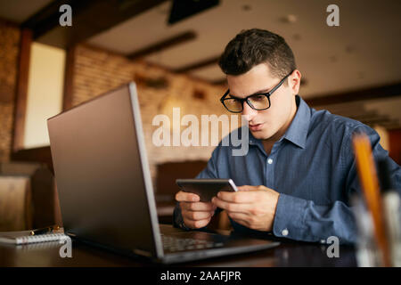 Handsome businessman distraite de travailler sur l'ordinateur portable regardant la vidéo sur smartphone. Freelancer holding mobile phone et l'aide de navigation 4g haute vitesse Banque D'Images