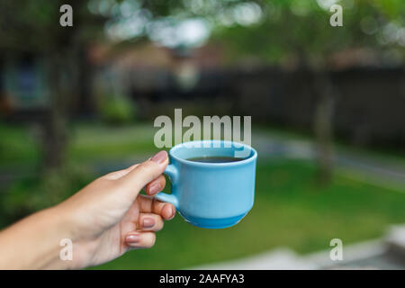 Gros plan, fille tient dans ses mains une tasse bleue avec un verre sur un arrière-plan flou d'un jardin. Banque D'Images