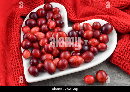 Canneberges rouges frais sur une plaque en forme de cœur entouré d'une serviette de cuisine rouge texturé. Concept pour le mois de la santé du cœur et de la Saint-Valentin. Banque D'Images