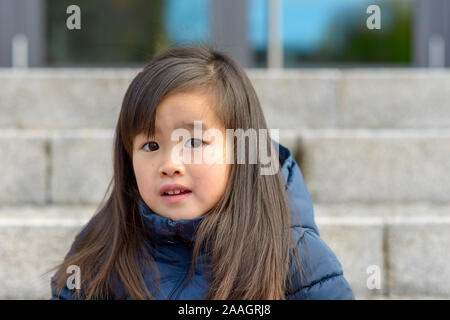 Pensive Young Asian girl à la peur qu'elle regarde la caméra avec parted lèvres tout en se posant à l'extérieur dans une veste chaude Banque D'Images