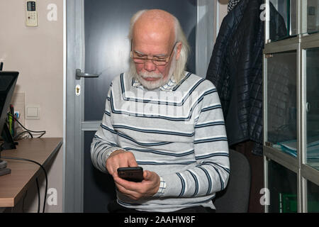 Close-up portrait demi-longueur du vieil homme chauve avec de longs cheveux gris et sa barbe en pullover rayé est l'utilisation de smartphone à l'intérieur du bureau. Banque D'Images