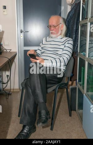 Close-up portrait de vieil homme chauve avec de longs cheveux gris et sa barbe en pullover rayé qui est à l'aide de smartphone dans l'intérieur du bureau. Banque D'Images