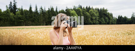 Voir l'échelle de l'image de jeune femme avec appareil photo reflex numérique professionnel prise de photos en extérieur dans la belle nature du champ de blé doré. Banque D'Images