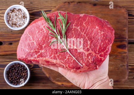 Man main tenant un steak au romarin frais crus au-dessus d'une tasse de fines herbes Banque D'Images